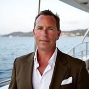A man in a brown blazer and white shirt standing on a boat with water and distant shoreline visible in the background